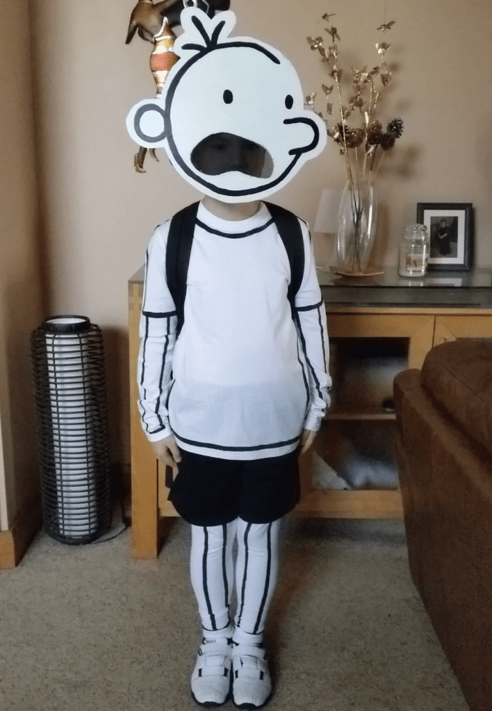 A person wearing a white shirt, black shorts, and white leggings is standing. They also have a cut out of a simply drawn face like the one the character in Diary of a Wimpy Kid has.