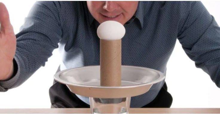 Egg on top of a toilet paper tube, standing on a plate on a glass of water, with a man ready to hit the plate (Eight Grade Science)