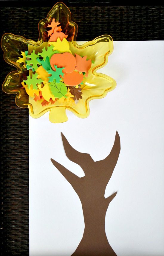 Fall art projects like this one show a piece of white paper with the outline of a tree in brown construction paper. a leaf shaped bowl holds a bunch of fall leaf stickers that are ready to be attached.