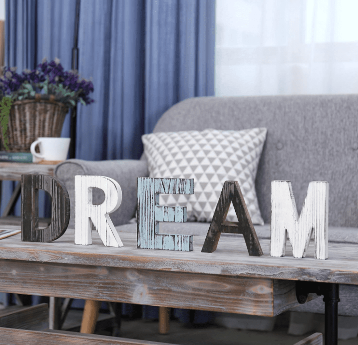 Multicolored and multitextured letters spell the word "DREAM" in all caps on a coffee table with a grey couch in the background.