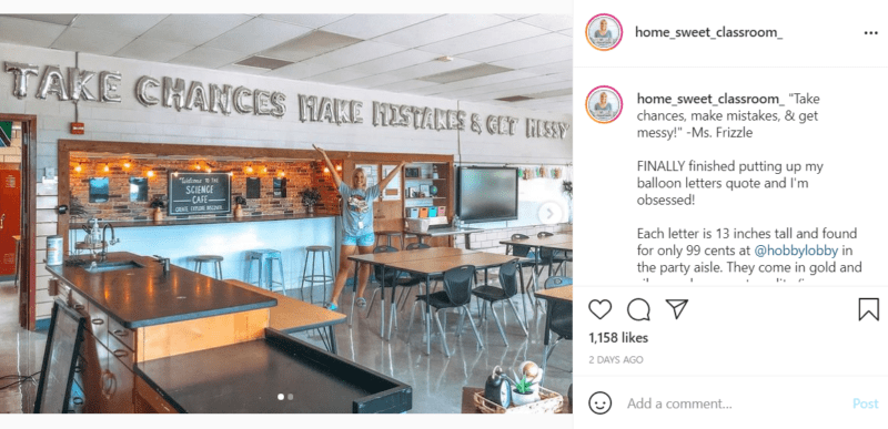 A science classroom with desks is displayed while a teacher holds up her arms towards silver balloons along the ceiling that say "TAKE CHANCES MAKE MISTAKES & GET MESSY."