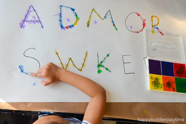 Child practicing writing letters by dipping fingertip in ink then pressing letters onto poster paper