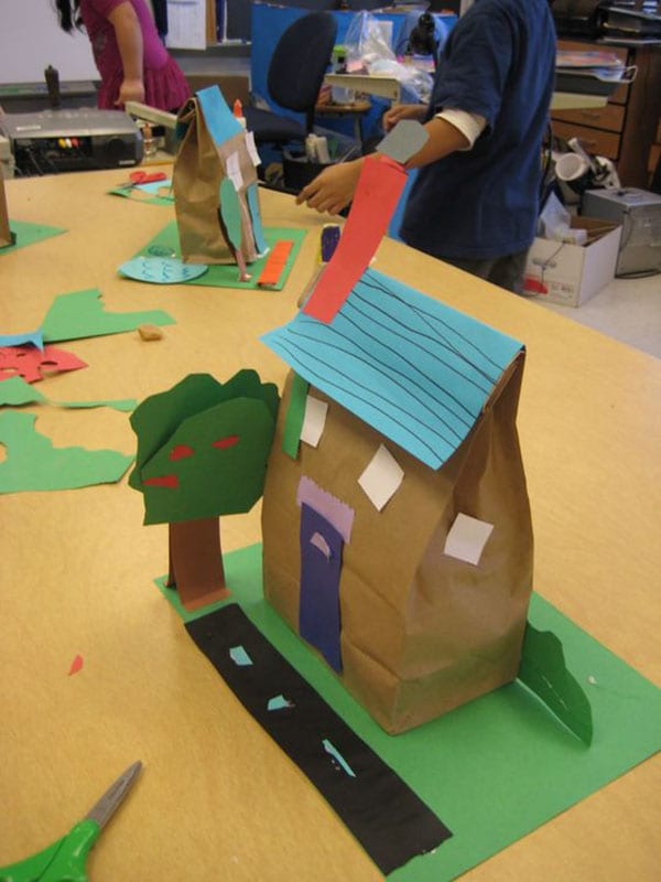 Paper lunch bag stuffed and turned into a house, sitting on construction paper lawn with a tree and road