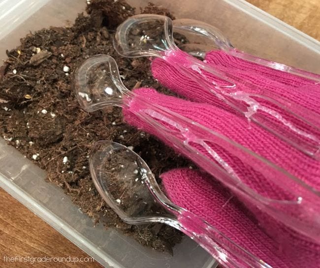 Student's hand wearing pink glove with plastic spoons attached to fingers, digging in dirt 