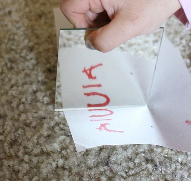Young student's hand holding a mirror on a piece of paper with the letters A I U, showing symmetry (First Grade Science Experiments)