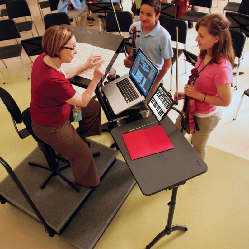 Music teacher in classroom seated at Flex Conductor's System Tech Bridge with laptop and speaking to two young students