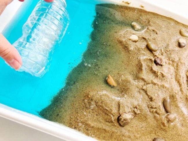 Cubo de plástico lleno de arena, conchas y agua para simular una playa, con una mano sosteniendo una botella de plástico en el agua (Ciencia de Cuarto grado)