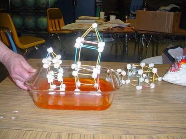 La mano dell'insegnante di scienze di quarta elementare che agita una padella di gelatina sormontata da un modello di casa fatto di stuzzicadenti e marshmallow's hand shaking a pan of Jello topped with a house model made of toothpicks and marshmallows