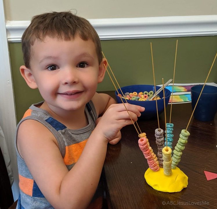 A little boy sits in front of a sculpture that has a base of play dough with raw spaghetti sticks coming out of it.  He is stringing fruit loops onto the spaghetti strands.  (fine motor activities)