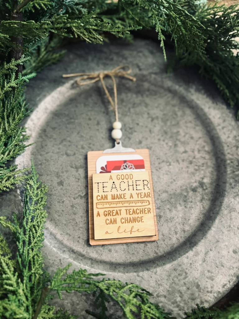 Teacher gift card holder that's also an ornament on a silver place with greenery.