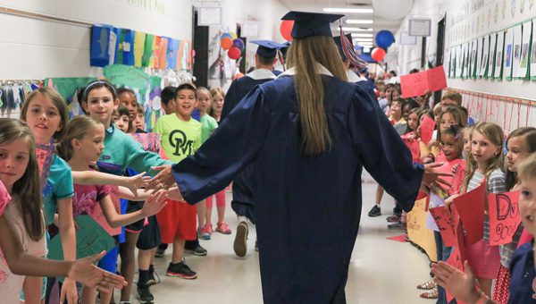 high school graduate in cap and gown walking hall of elementary school giving students high fives