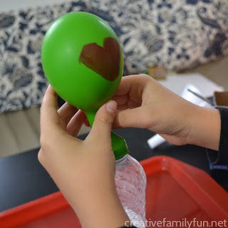 Hands are shown holding a green balloon that has a heart drawn on it. The balloon is partially filled. 
