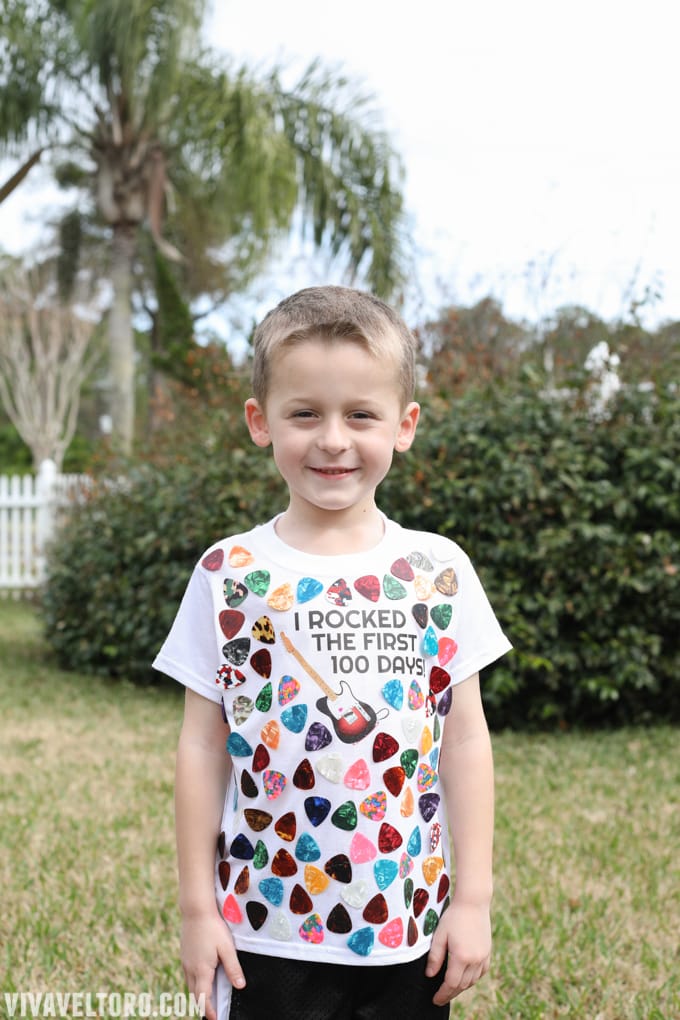 A little boy is shown wearing a t-shirt with guitar picks all over it. It says I Rocked the First 100 Days! (100th day of school shirt ideas)