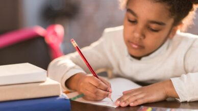 african american child writing in a notebook at a desk with stacks of book