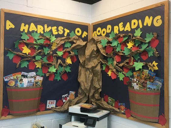 October bulletin boards can cover two walls like this one. It reads "A harvest of good reading." A tree with fall leaves spreads out across the two walls. Books are in two barrels at the bottom of each wall. 