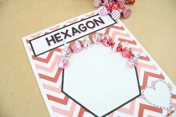 Hexagon is written across the top of a mat and a hexagon shape is shown surrounded by pink and red lines. Valentine's Day hershey kisses are shown lining the shape. (Valentine's Day Crafts for Preschoolers)