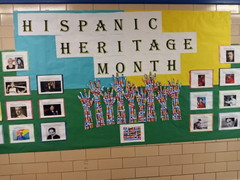 A bulletin board reads Hispanic Heritage Month in black letters on a white background. Ten hands are created from a number of different flags. There are several photos of famous people of Hispanic descent. 
