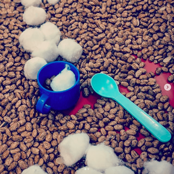 Beans, play mugs, spoons, and cotton balls in a sensory table for classroom pretend play making hot cocoa
