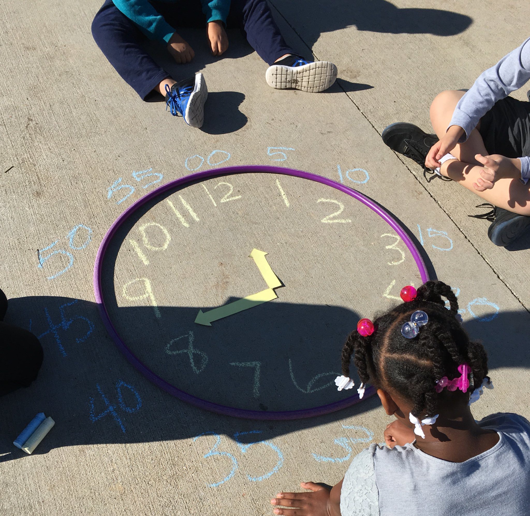 Kids are shown sitting on the ground. A purple hula hoop creates the perimeter of a clock and the numbers have been written in chalk.