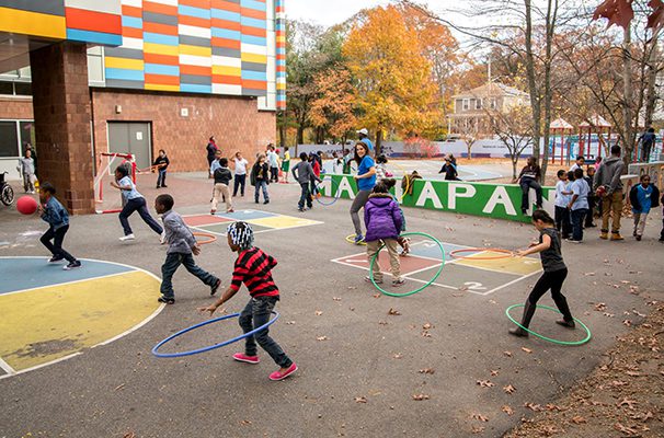 Students are shown on an outdoor courtyard with hula hoops around their feet and legs demonstrating one of many recess games that can be played with hula hoops.