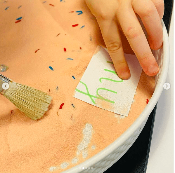 Pink salt in a tray with colored sprinkles and a child's hand holding a paintbrush to brush salt off a card with the sight word "my"