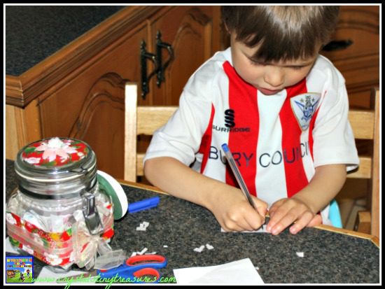 A child sits in a chair beside a highly decorated jar writing on a strip of paper.