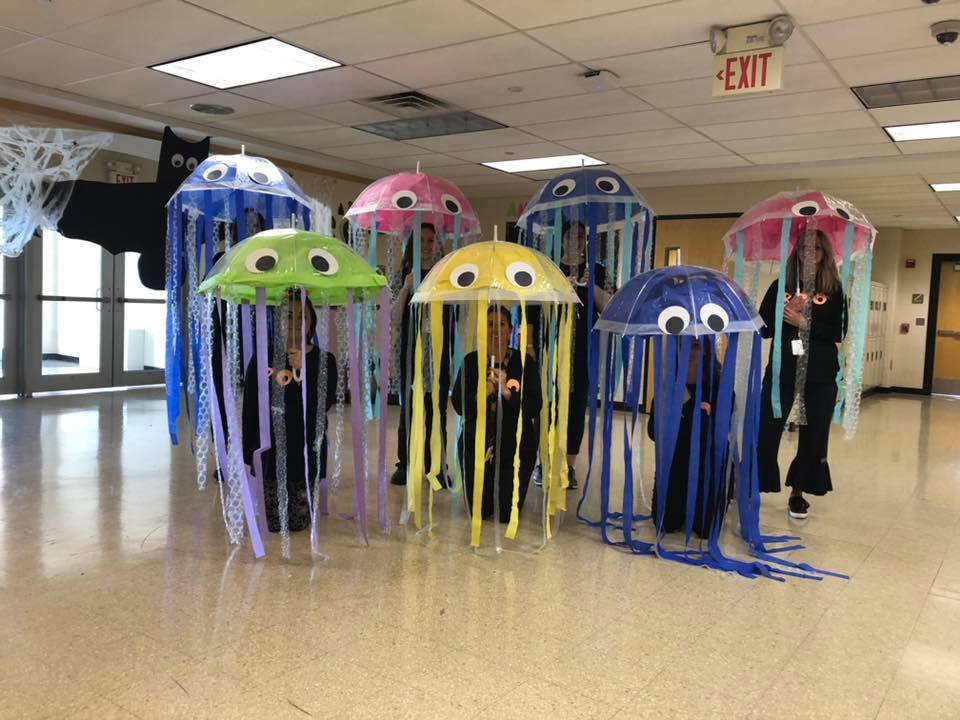 7 teachers stand under different colored umbrellas that have eyes attached to make them look like jellfish. There are streamers coming down from each umbrella.