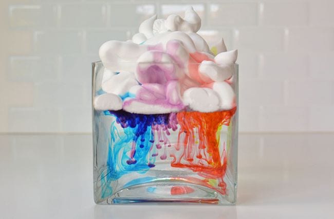 Clear jar filled with water, with shaving cream floating on top and water coloring dripping from the shaving cream, as an example of kindergarten science activities