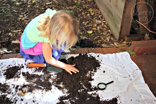 Child examining dirt on a white tarp with a magnifying glass (Kindergarten Science Activities)