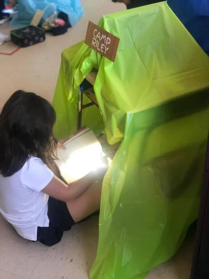 A little girl is sitting by a makeshift tent reading by lantern light.