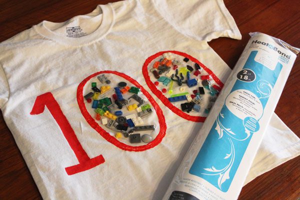 A white t-shirt has a big 100 in red on it. Inside the zeros are legos that have been glued down.