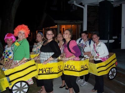 Eight women are inside a cardboard bus that reads Magic School Bus. The woman in the front has a red, curly wig on. 