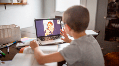 Young boy sitting at messy desk waving to teacher on laptop screen for online tutoring