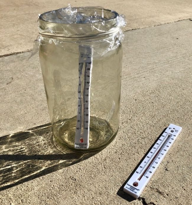 Glass jar covered in plastic wrap with a thermometer inside, next to a thermometer lying on the ground (Outdoor Science)
