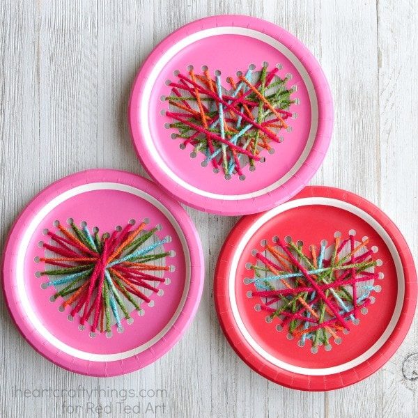 Three pink and red paper plates are shown with hole punches in the shape of hearts on them.  They have strong yarn between the holes.