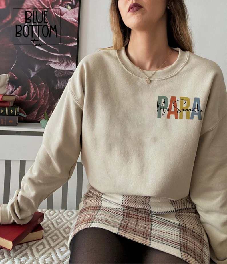 A woman is shown from the neck down.  She is wearing an off white crewneck sweatshirt that says PARA and her name on it.  (gifts for paraprofessionals)