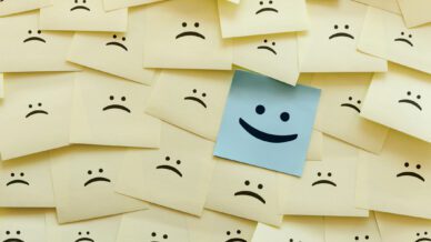 One blue smiley face sticky note on background of yellow frowny face sticky notes