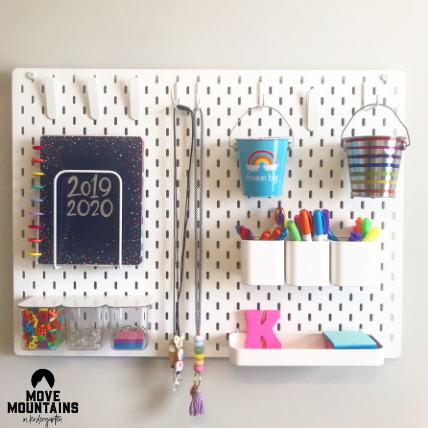 A white pegboard is shown with some containers clipped onto it with art supplies and other school supplies.