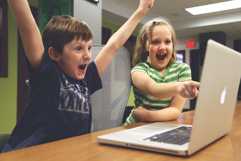 Un niño levanta los brazos en señal de victoria mientras una niña señala la pantalla de una computadora y sonríe (gesto de escritura de imágenes)