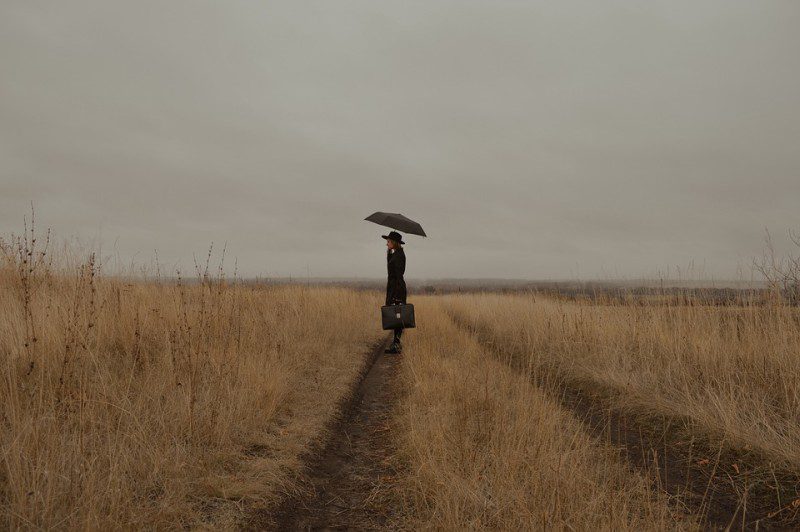 Una mujer parada en medio de un campo de trigo en un día gris, cargando un paraguas y una bolsa.