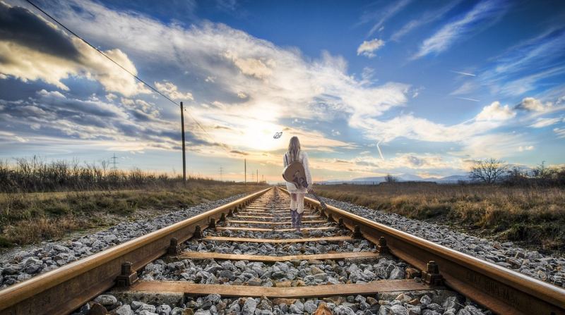 Mujer de pie sobre las vías del tren sosteniendo la guitarra y mirando la puesta de sol (leyenda de la imagen)