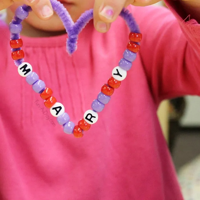 Hands are shown holding a heart made from a purple pipe cleaner. It has beads on it and some of them spell Mary (Valentine's Day Crafts for Preschoolers)