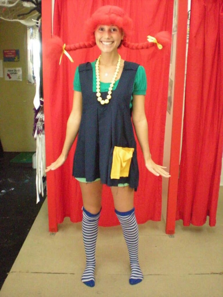Book character costume ideas like this one shows a woman dressed as Pippi Longstocking. She has one a blue overall style dress, knee socks, and the signature red braided wig and the braids are sticking out on edge.