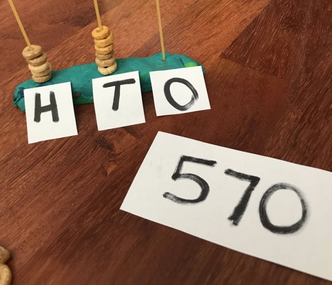  stranks spaghetti straight upright into playdough and labeled H, T, and O with Cheerios stacked on each next to card reading 570