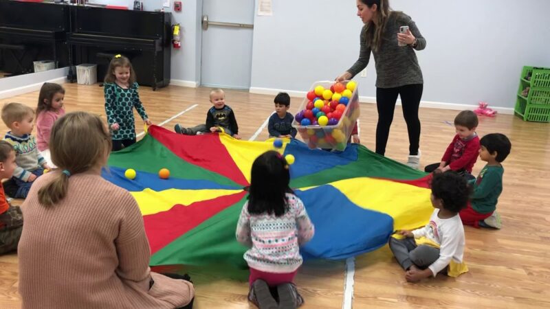Several small children and a few adults sit around a multi-colored parachute that has colorful balls being dumped on it as part of recess games..