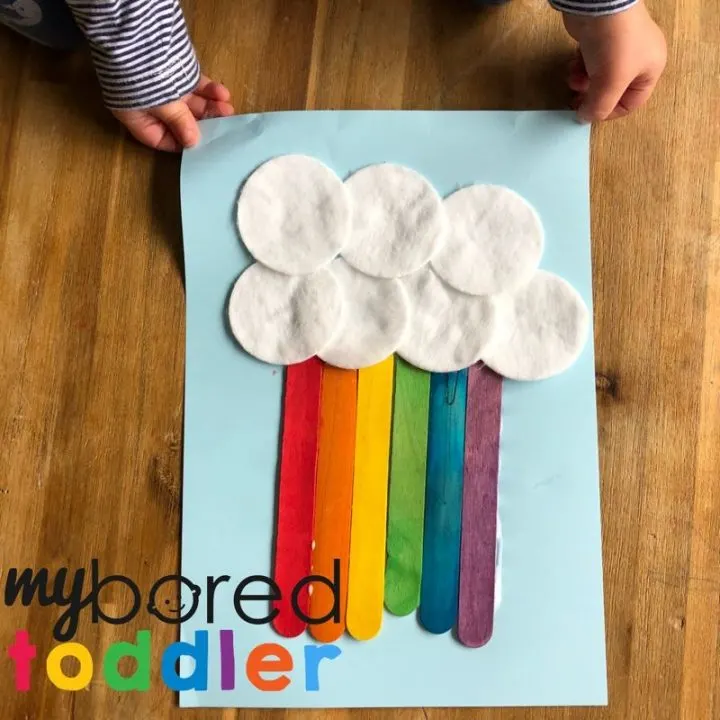 This easy art project for kids shows white circles glued to a piece of light blue construction paper as clouds. Red, orange, yellow, green, blue, and purple popsicle sticks are coming out of the clouds as a rainbow.