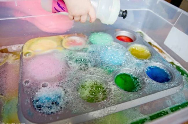 Preschool student squeezing a bottle of vinegar into muffin tin cups filled with baking soda and food coloring