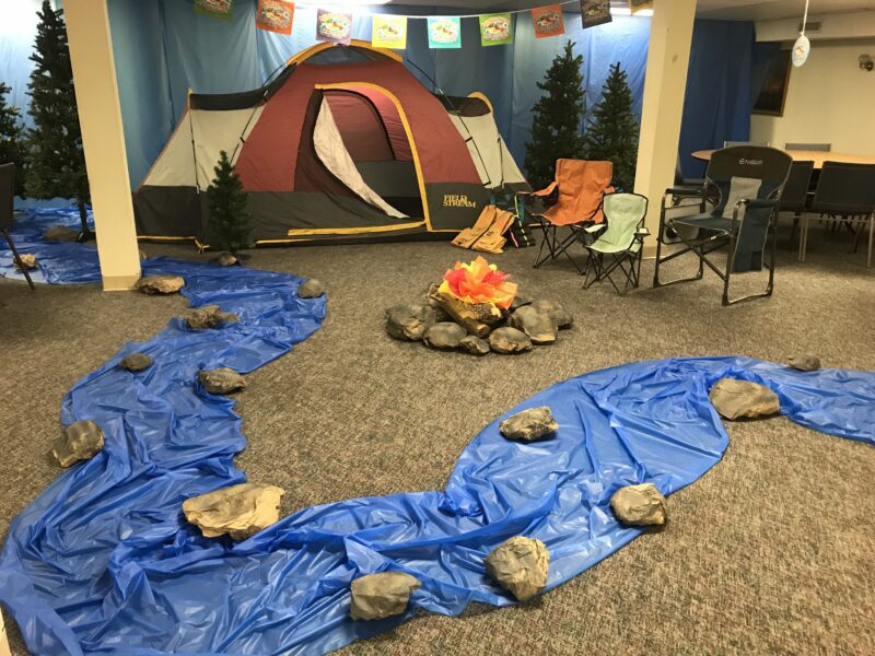 A pretend river is made from blue plastic tablecloths with large rocks spread throughout. A tent and a pretend campfire are also shown. 