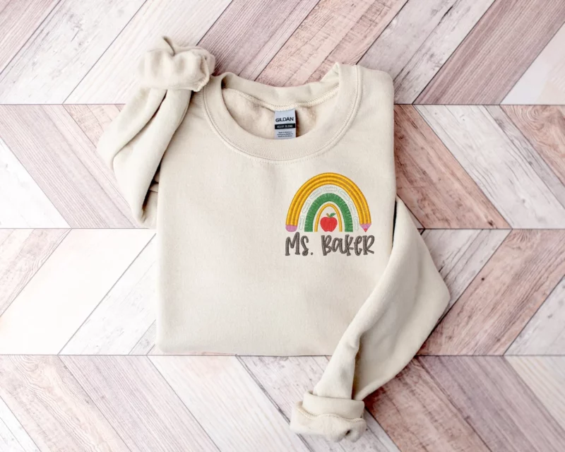 A beige sweatshirt has a rainbow in the corner with a teacher's name embroidered on it.