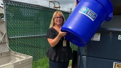 A woman dumping a blue recycling trash can into a dumpster